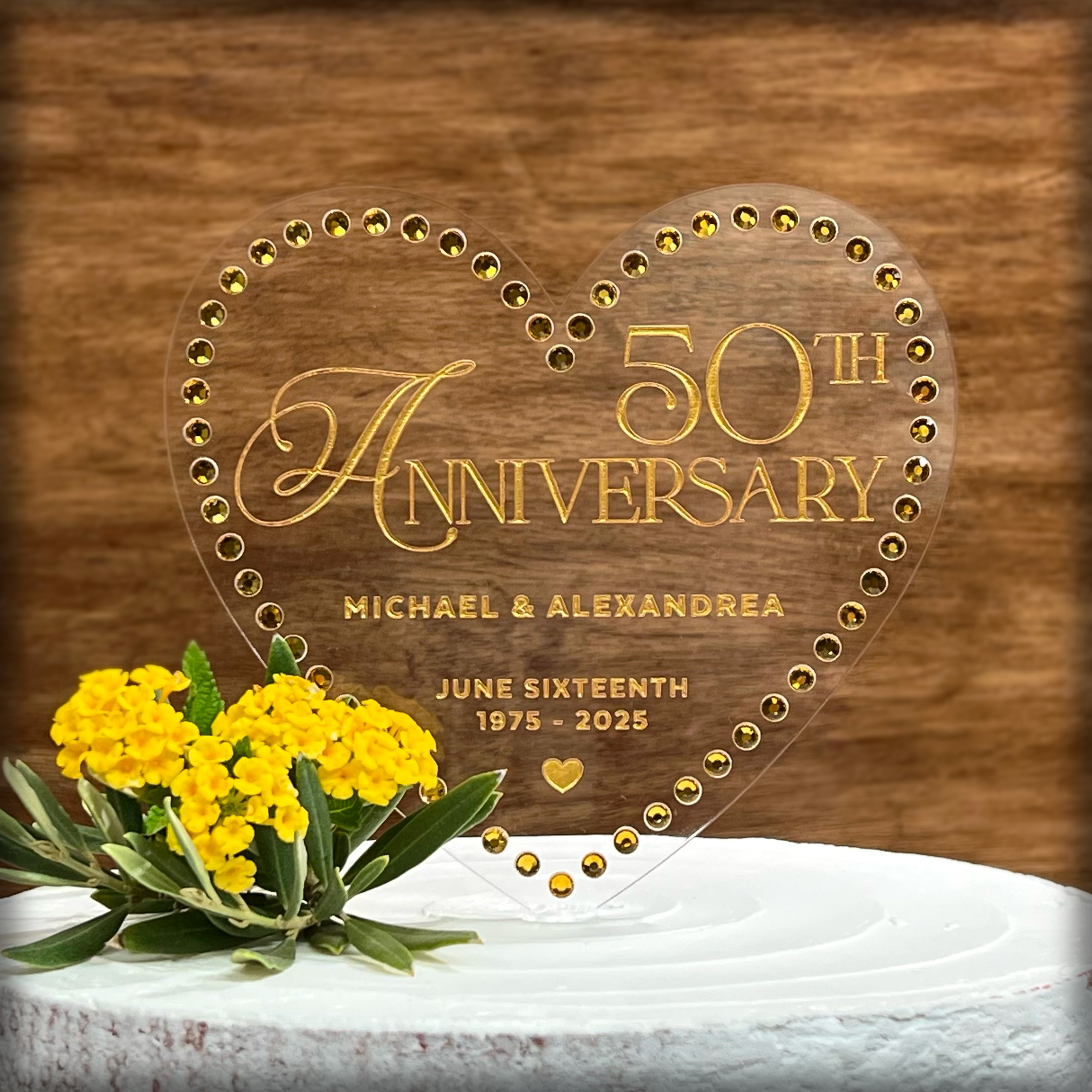 A classic symbol of love, the heart shape makes it the perfect choice for an anniversary cake topper. The clear acrylic material gives the topper a modern and sleek look that will complement any cake design. The gold rhinestone border and gold painted text, which reads "Happy Anniversary", adds a touch of glamour and luxury to the topper, making it stand out as a true centerpiece of your anniversary celebration capturing the essence of the occasion. This anniversary cake topper is not only beautiful, but a long-lasting cherished keepsake for years to come.